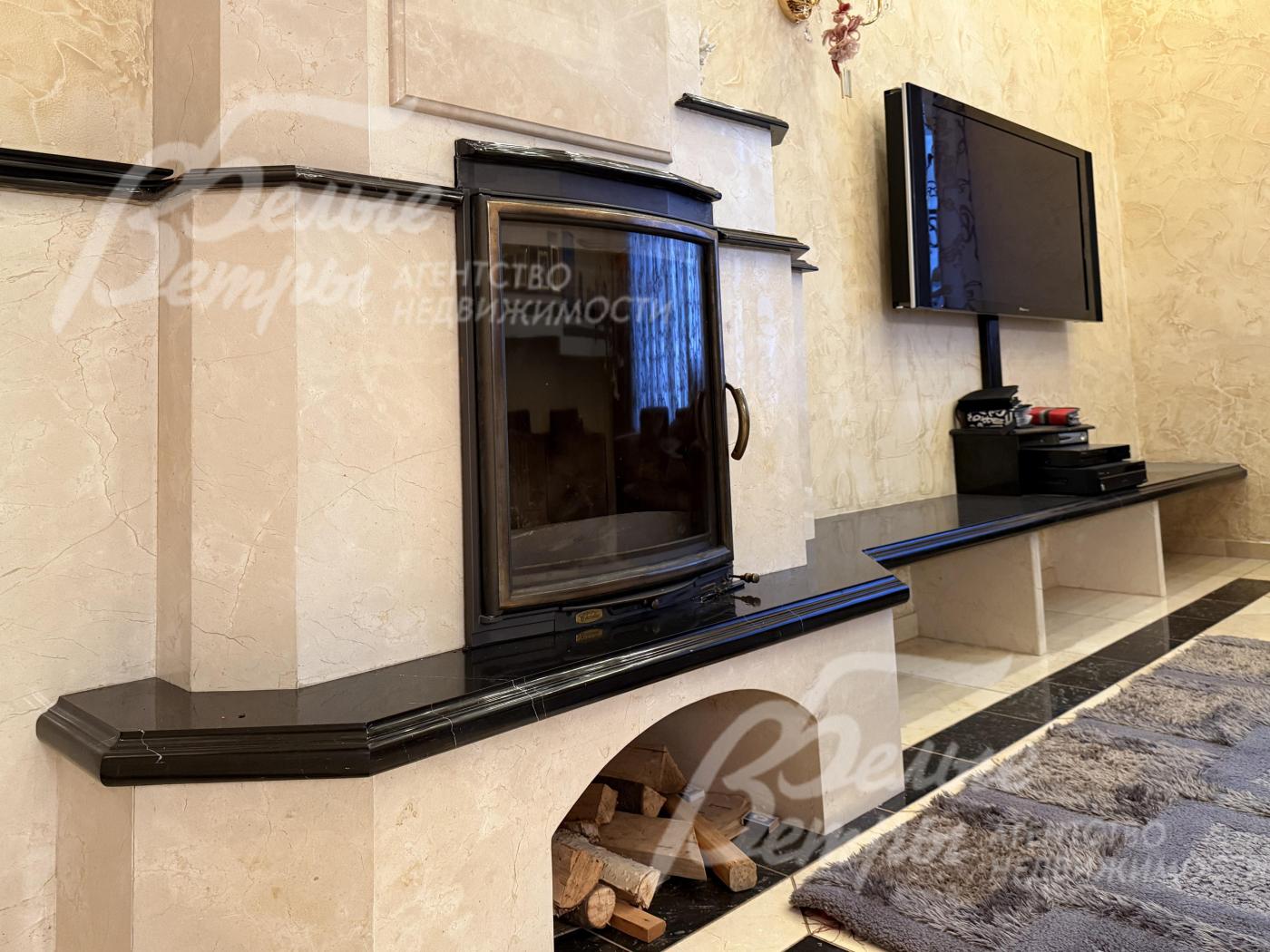 Камин - со стеклянной дверцей, облицовка выполнена из светлого камня с нежной текстурой и контрастного тёмного камня, под топкой - ниша для хранения дров