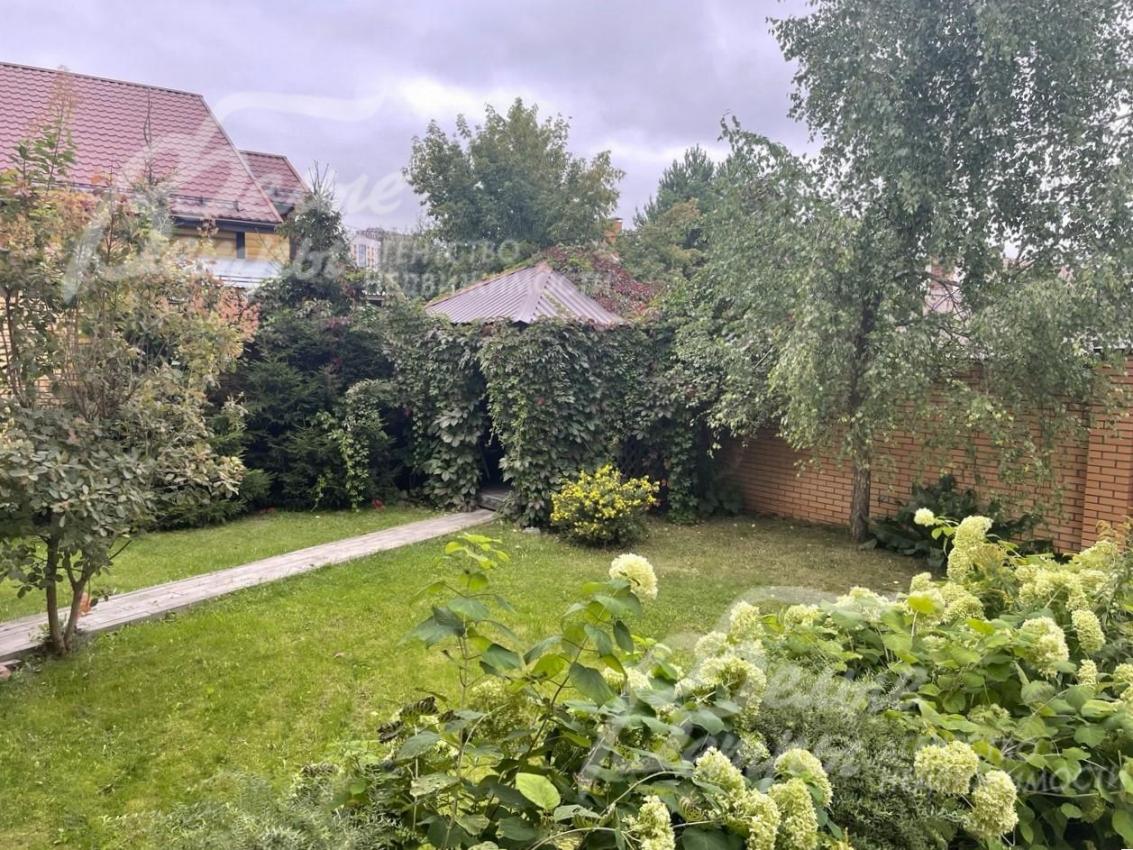 На участке крытая беседка и множество цветущих растений.